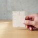 Lit 160×200 à l’échelle 1/25 tenu en main – Kit Yoja Main tenant un lit en carton du Kit Yoja à l’échelle 1/25 (6,4×8 cm) sur bureau en bois clair avec fond béton flouté et lumière naturelle